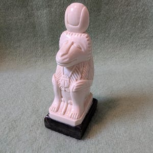 Puede incluir: Una figura tallada de color crema de un babuino sentado con un tocado redondeado, con texturas de pelaje detalladas y un collar, montada sobre una base rectangular negra. La escultura representa al animal en una postura formal y erguida.