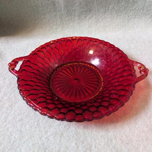 Puede incluir: Un cuenco redondo de cristal rojo rubí con un patrón de panal texturizado en toda su superficie y un diseño de rayos de sol en el centro. El cuenco incluye dos pequeñas asas integradas en los lados opuestos y un borde festoneado.