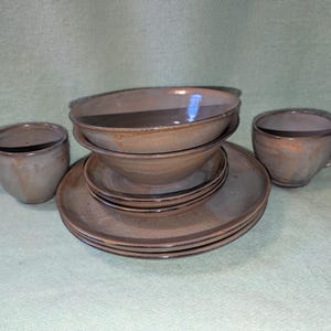 Può includere: Un set di stoviglie in ceramica composto da due tazze, due ciotole e due piatti. I pezzi presentano una finitura liscia e lucida in toni tenui di marrone e grigio con bordi più scuri e leggermente irregolari. Gli articoli sono impilati.
