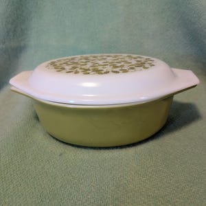 May include: A round, light green casserole dish with a matching white lid. The lid features a repeating pattern of small, olive green leaves and white berries. The dish has two integrated side handles and a smooth, opaque glass construction.
