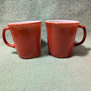Peut inclure: Deux tasses assorties avec une finition extérieure lisse et unie orange brûlé et un intérieur de couleur claire contrastante. Chaque tasse présente un corps arrondi, une anse latérale robuste et un bord légèrement évasé au design épuré.