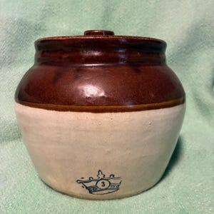 Peut inclure: Un pot en grès avec un corps arrondi et un couvercle. La partie supérieure est vernissée en brun foncé, tandis que la partie inférieure est de couleur crème. Un motif bleu estampillé représentant une couronne avec le chiffre 3 est visible.