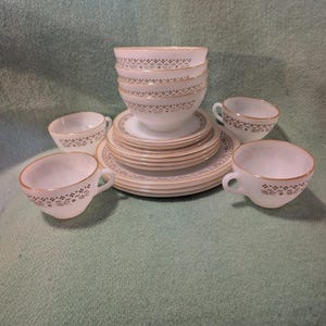 Könnte beinhalten: Ein Set aus weißem Milchglas-Geschirr mit einem zarten, goldfarbenen Blumen- und Geometriemuster am Rand. Die Kollektion umfasst einen Stapel Schalen, mehrere flache Teller und vier passende Teetassen mit Henkeln, alle mit einheitlichem Dekor.