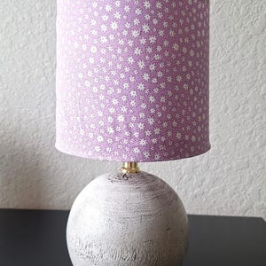 May include: A table lamp featuring a spherical base with a distressed white wood grain finish and a cylindrical lampshade in a light purple fabric adorned with a repeating pattern of small white star-shaped flowers and a gold-tone metal neck.