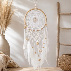 Può includere: Un acchiappasogni circolare con un cerchio in legno chiaro e un'intricata trama in macramè bianco. Il design include cordoncini di cotone bianco, perline di legno lisce e diverse piume bianche morbide sospese nella parte inferiore.