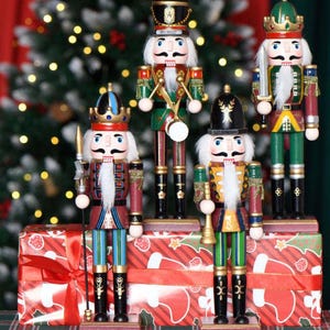 Puede incluir: Cuatro figuras de cascanueces de madera sobre una caja de regalo roja con patrones de calcetines blancos. Las figuras visten uniformes coloridos en rojo, verde, azul y dorado, con coronas o sombreros, barbas de piel sintética y accesorios.