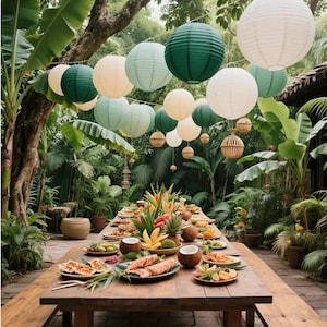 May include: A long wooden dining table set outdoors, laden with plates of tropical fruit, sliced coconuts, and seafood. Above the table, numerous round paper lanterns in shades of white, light green, and dark green hang from strings in the trees.