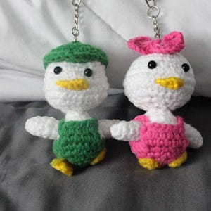Peut inclure: Deux porte-clés canards en crochet avec des fermoirs mousquetons en métal argenté. Un canard porte un chapeau et une salopette verts, l'autre un nœud et une salopette roses. Ils ont des corps en laine blanche, des yeux noirs et des becs jaunes.