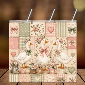 Könnte beinhalten: Ein zylindrischer Becher mit Strohhalm und einem Patchwork-Design aus zartrosa, salbeigrünen und cremefarbenen Quadraten. Das Muster zeigt drei weiße Gänse mit Blumenkränzen und Schleifen, umgeben von Herzen, Schleifen und Blumen.