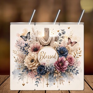 Könnte beinhalten: Ein zylindrischer Becher mit einem Aquarellmotiv, das ein mit Stoff drapiertes Holzkreuz zeigt, umgeben von blauen, rosafarbenen und cremefarbenen Blumen und Schmetterlingen. Ein zentrales Emblem zeigt das Wort Blessed in goldener Schrift.