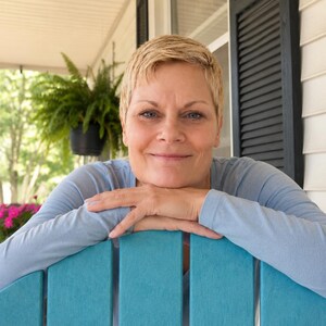 May include: A person with short blonde hair wearing a light blue long-sleeve shirt, resting their chin on their hands while leaning against the back of a bright teal wooden chair with visible silver screws on the vertical slats.
