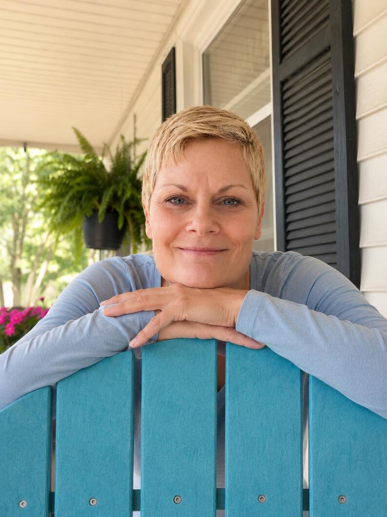 May include: A person with short blonde hair wearing a light blue long-sleeve shirt, resting their chin on their hands while leaning against the back of a bright teal wooden chair with visible silver screws on the vertical slats.