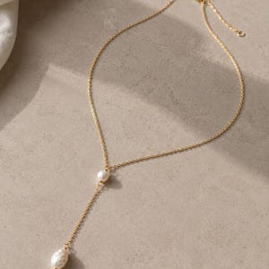 Puede incluir: Un collar de cadena en tono dorado con diseño de lazo, que presenta una pequeña perla blanca redonda situada en la unión central y un colgante de perla blanca de forma irregular que cuelga del extremo del segmento de cadena vertical.