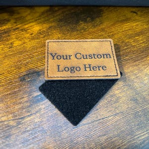 May include: A rectangular tan leather patch with stitched edges featuring the black engraved text Your Custom Logo Here. The patch is attached to a black hook-and-loop fastener backing, creating a functional accessory for gear or apparel.