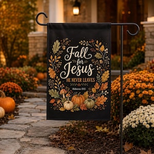 Puede incluir: Una bandera de jardín rectangular negra con el texto Fall for Jesus HE NEVER LEAVES Hebrews 13:5 en letras blancas y doradas. El diseño está rodeado por una colorida corona otoñal de hojas naranjas, amarillas y verdes, bellotas y calabazas.