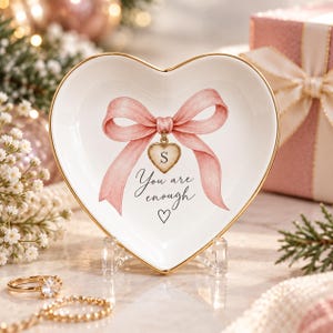 Könnte beinhalten: Eine herzförmige Schmuckschale aus weißer Keramik mit goldfarbenem Rand. In der Mitte befindet sich eine rosa Schleifenillustration, ein kleiner herzförmiger Anhänger mit dem Buchstaben S und der schwarze Text "You are enough" mit einem Herz.