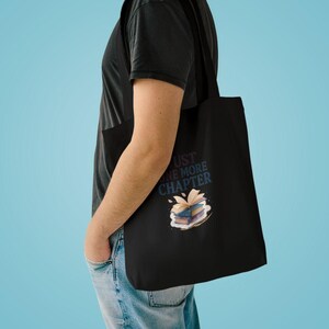 Op de afbeelding: Een zwarte tote bag met een afbeelding van een stapel boeken met een open boek erbovenop. De tekst "JUST ONE MORE CHAPTER" is in het blauw boven de illustratie gedrukt. De tas heeft twee schouderbanden en is gemaakt van zwarte stof.