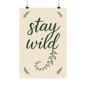 May include: A rectangular wall poster with a cream-coloured background featuring the dark green text stay wild in a cursive script. The letter d extends into a leafy vine graphic, and four small fern-like sprigs are positioned in the corners.