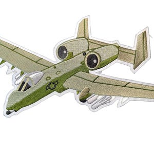 May include: An embroidered patch of an A-10 Thunderbolt II military aircraft in olive green and tan colors. The design features a detailed cockpit, two large jet engines mounted above the wings, a twin tail configuration, and visible underwing weaponry.