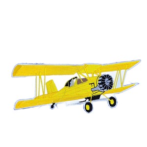 May include: A bright yellow biplane embroidered patch featuring two stacked wings, a dark gray radial engine, a small cockpit area, and black landing gear wheels. The design is rendered in detailed yellow thread with visible stitching throughout.