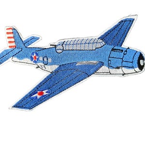 May include: An embroidered patch shaped like a blue military aircraft. The plane features a white underside, a red and white striped tail fin, and circular insignia with a red star and blue accents on the fuselage and the wing.