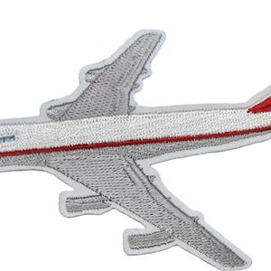May include: An embroidered iron-on patch shaped like a commercial airplane. The aircraft is primarily silver and white with a thin red stripe running along the fuselage. The tail fin features a red accent and the text 747 in black lettering.