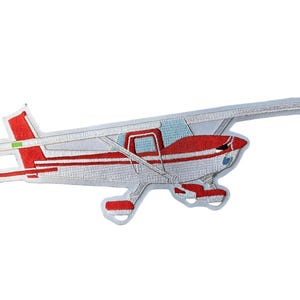May include: An embroidered patch shaped like a small propeller airplane. The aircraft features a white body with red stripes, a red tail section, and light blue windows. The design includes a high-wing configuration and landing gear wheels.