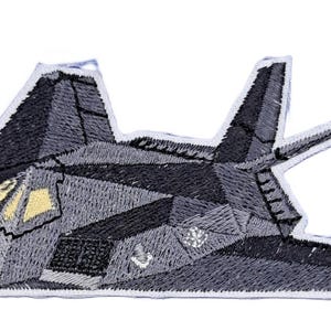 May include: An embroidered patch depicting a dark gray stealth fighter jet in profile. The design features angular, geometric shapes with a light yellow cockpit window and white stitched outlines, showcasing a detailed, textured thread construction.
