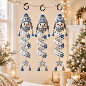 Puede incluir: Tres adornos colgantes de muñeco de nieve con cabezas tejidas a ganchillo y gorros de invierno con patrones en azul y gris. Cada cuerpo es una cinta blanca en espiral decorada con copos de nieve azules y estrellas, terminada en una estrella plateada.