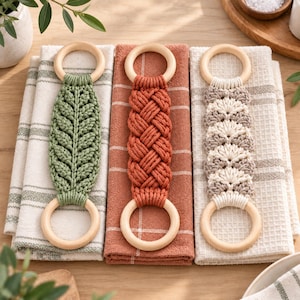 Könnte beinhalten: Drei Makramee-Handtuchhalter mit hellen Holzringen an beiden Enden. Der linke Halter ist salbeigrün mit Blattmuster, der mittlere ist terrakottafarben mit Flechtmuster und der rechte ist cremefarben und beige mit einem Muschelmuster.