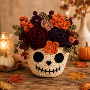 Könnte beinhalten: Ein cremefarbener, gehäkelter Totenkopf-Übertopf, gefüllt mit einem lebendigen Arrangement aus gehäkelten Blumen und Beeren in den Farben Dunkelburgunder, Dunkellila, leuchtendes Orange und Olivgrün, mit schwarzen gestickten Augen, Nase und Mund.