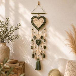 Puede incluir: Un tapiz de pared con un gran corazón central de ganchillo en color crema y verde oscuro, suspendido de un listón de madera. Debajo, tres corazones más pequeños y detalles en forma de hoja cuelgan de hilos con cuentas de madera y borlas.