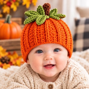 May include: A bright orange crochet hat designed to resemble a pumpkin, featuring vertical ribbed texture, a cluster of green crocheted leaves on top, and a small brown crocheted stem centered among the leaves, worn by a smiling infant.