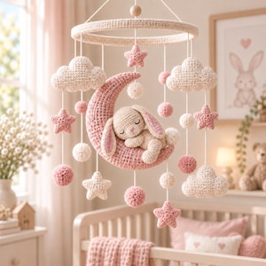 Può includere: Una giostrina per culla all'uncinetto con una mezzaluna rosa centrale e un coniglietto color crema che dorme. La giostrina include nuvole, stelle e piccoli pon pon all'uncinetto color crema e rosa appesi a una cornice con fili bianchi.