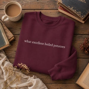 May include: A deep burgundy crewneck sweatshirt featuring the white text "what excellent boiled potatoes" printed across the centre of the chest. The garment has long sleeves, a ribbed neckline, and a simple, clean design on the front.