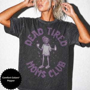 May include: A charcoal gray short-sleeve t-shirt featuring a purple graphic of a skeleton wearing a t-shirt and slippers, holding a mug and a baby bottle. The text "DEAD TIRED MOMS CLUB" is printed in a purple arched font around the skeleton.