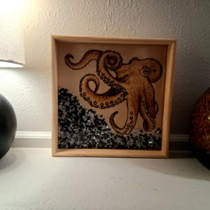 May include: A square wooden frame containing a tan octopus illustration on a light background. The bottom section of the frame is filled with small, dark gray and black stones, creating a textured, layered appearance beneath the octopus artwork.