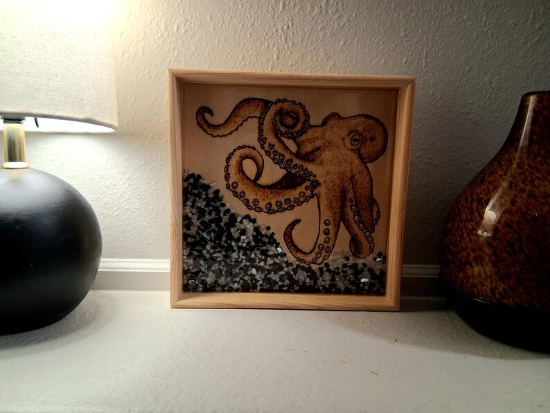 May include: A square wooden frame containing a tan octopus illustration on a light background. The bottom section of the frame is filled with small, dark gray and black stones, creating a textured, layered appearance beneath the octopus artwork.