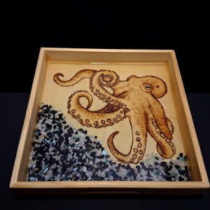 May include: A square wooden tray featuring a wood-burned octopus illustration on a light tan base. The bottom corner is filled with a layer of small, dark blue, black, and clear glass pebbles embedded in a smooth, glossy resin finish.