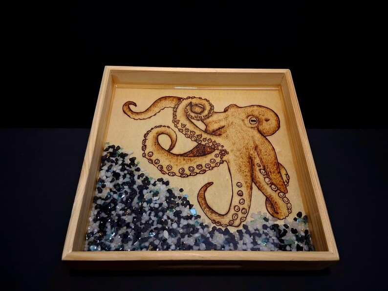 May include: A square wooden tray featuring a wood-burned octopus illustration on a light tan base. The bottom corner is filled with a layer of small, dark blue, black, and clear glass pebbles embedded in a smooth, glossy resin finish.