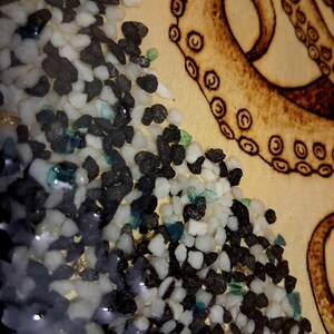 May include: A light-colored wooden surface featuring a wood-burned octopus tentacle with visible suction cups. Adjacent to the tentacle is a textured section filled with small, loose black and white stones mixed with tiny flecks of blue and gold.