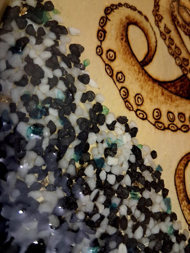 May include: A light-colored wooden surface featuring a wood-burned octopus tentacle with visible suction cups. Adjacent to the tentacle is a textured section filled with small, loose black and white stones mixed with tiny flecks of blue and gold.