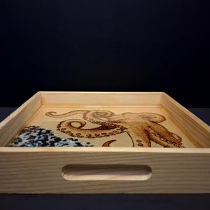 May include: A rectangular light-colored wooden serving tray with cutout handles on the sides. The base features a detailed wood-burned illustration of an octopus and a section filled with small, dark blue and white decorative stone fragments.