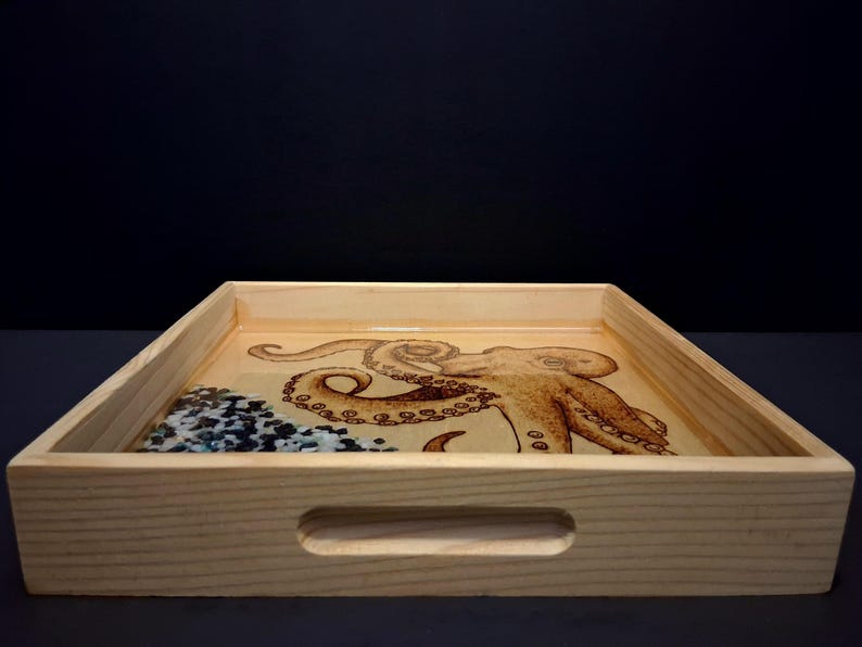 May include: A rectangular light-colored wooden serving tray with cutout handles on the sides. The base features a detailed wood-burned illustration of an octopus and a section filled with small, dark blue and white decorative stone fragments.
