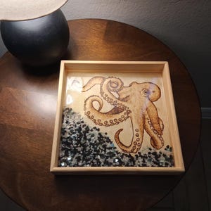 May include: A square wooden tray featuring a detailed illustration of a tan octopus with curled tentacles. The bottom section of the tray is filled with a layer of small, polished black and white stones embedded in a clear, glossy resin finish.