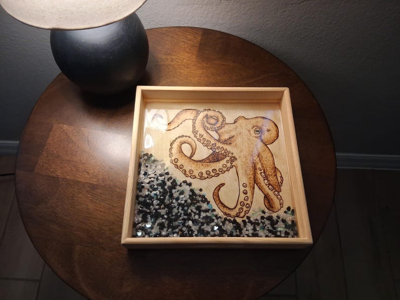 May include: A square wooden tray featuring a detailed illustration of a tan octopus with curled tentacles. The bottom section of the tray is filled with a layer of small, polished black and white stones embedded in a clear, glossy resin finish.