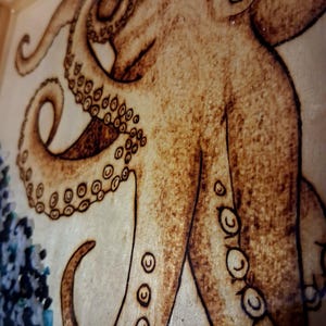 May include: A wood-burned artwork on a light-colored wooden surface featuring a detailed octopus with prominent suckers on its tentacles. The octopus is rendered in shades of brown, and a cluster of small, dark blue and black stones is visible.