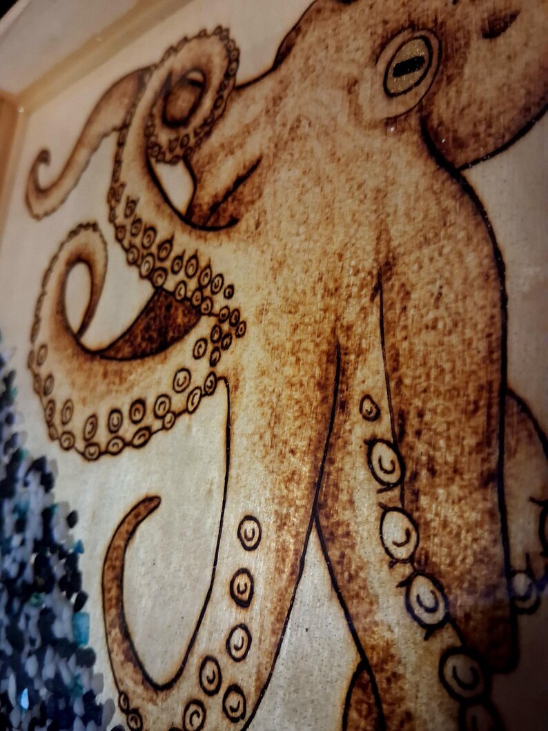 May include: A wood-burned artwork on a light-colored wooden surface featuring a detailed octopus with prominent suckers on its tentacles. The octopus is rendered in shades of brown, and a cluster of small, dark blue and black stones is visible.