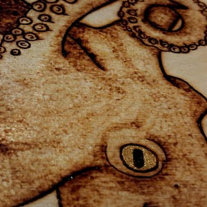May include: A wood-burned illustration of an octopus on a light-colored wooden surface. The design features dark brown scorched lines detailing the tentacles with circular suckers and a prominent eye with a gold-colored, glittery iris.