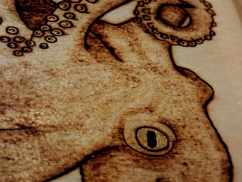 May include: A wood-burned illustration of an octopus on a light-colored wooden surface. The design features dark brown scorched lines detailing the tentacles with circular suckers and a prominent eye with a gold-colored, glittery iris.